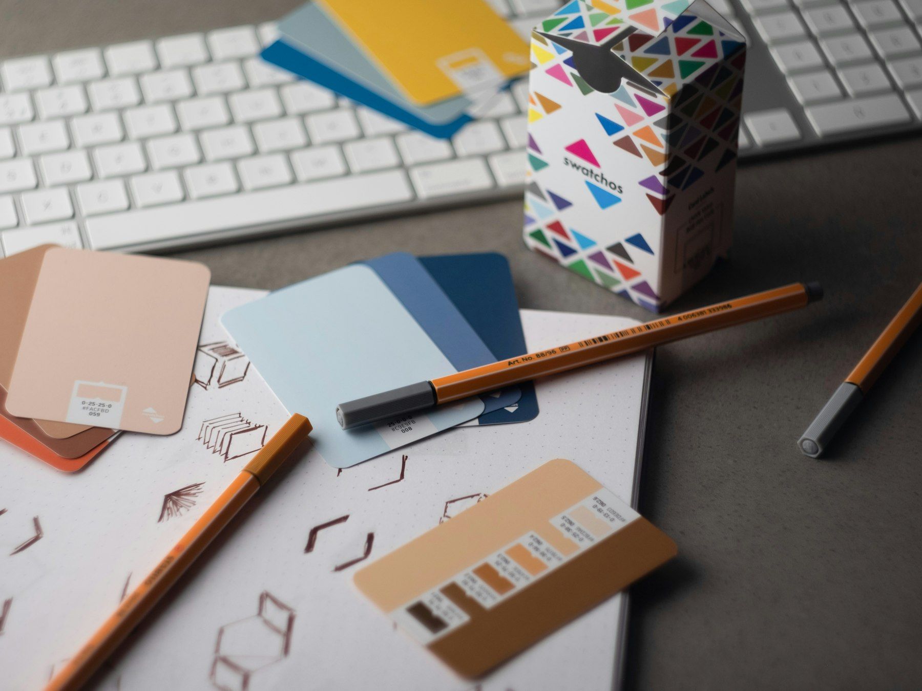 Colour swatch cards, pens and a sketchbook on a desk beside a keyboard